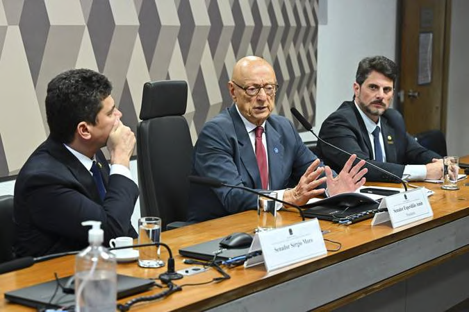 Três participantes à mesa, com um deles falando ao microfone.